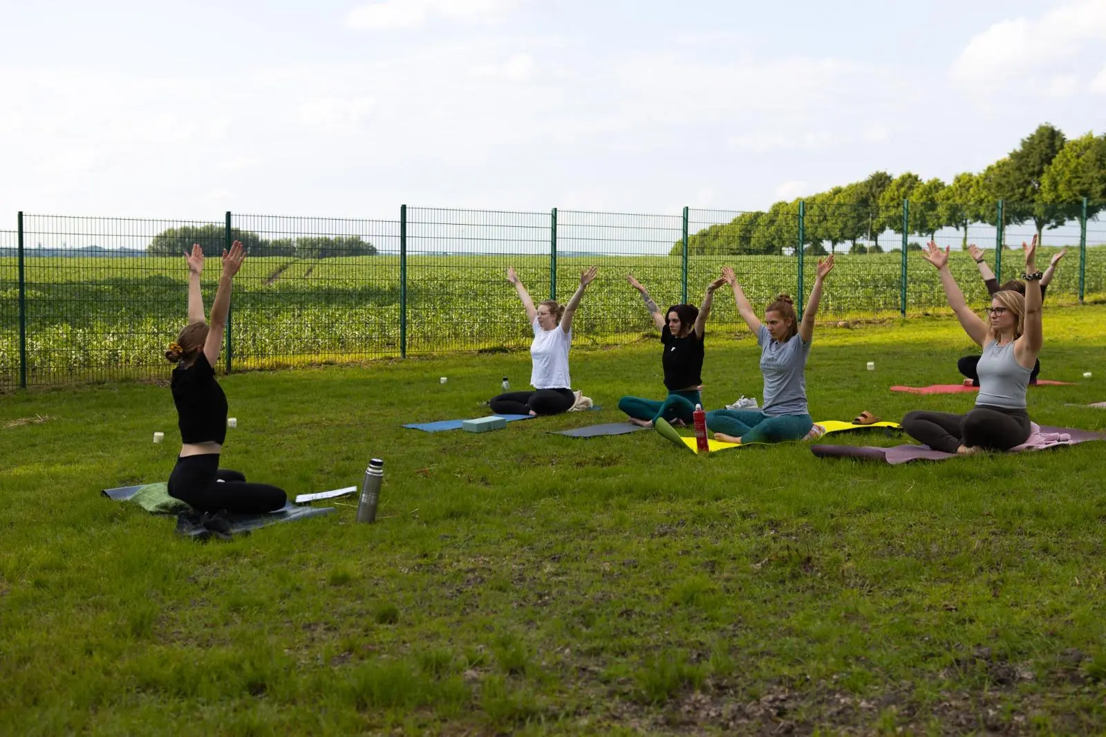 Yogagruppe in NRW auf der Wiese – sitzende Position mit erhobenen Armen, umgeben von Feld und Bäumen